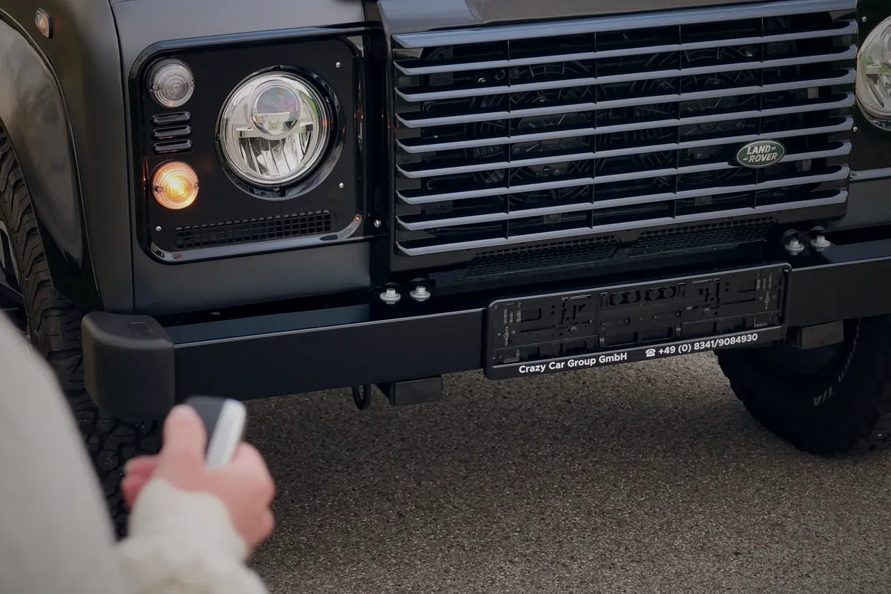 Hand mit Fahrzeugschlüssel vor der Front eines dunklen Land Rover Defender, der Blinker leuchtet beim Entriegeln