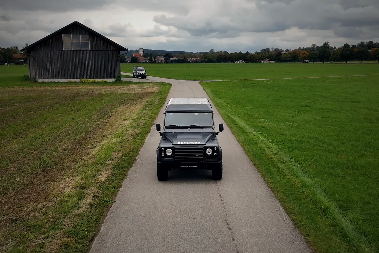 Dunkelgrauer Land Rover Defender auf einem schmalen Feldweg, dahinter ein Holzstadel und ein Dorf mit Kirchturm