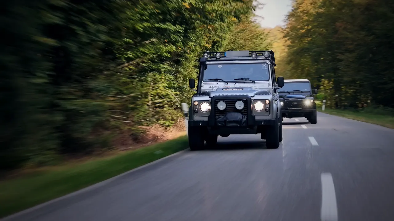 Silberner Land Rover Defender mit eingeschaltetem Licht auf einer Waldstraße, dahinter ein schwarzer Defender